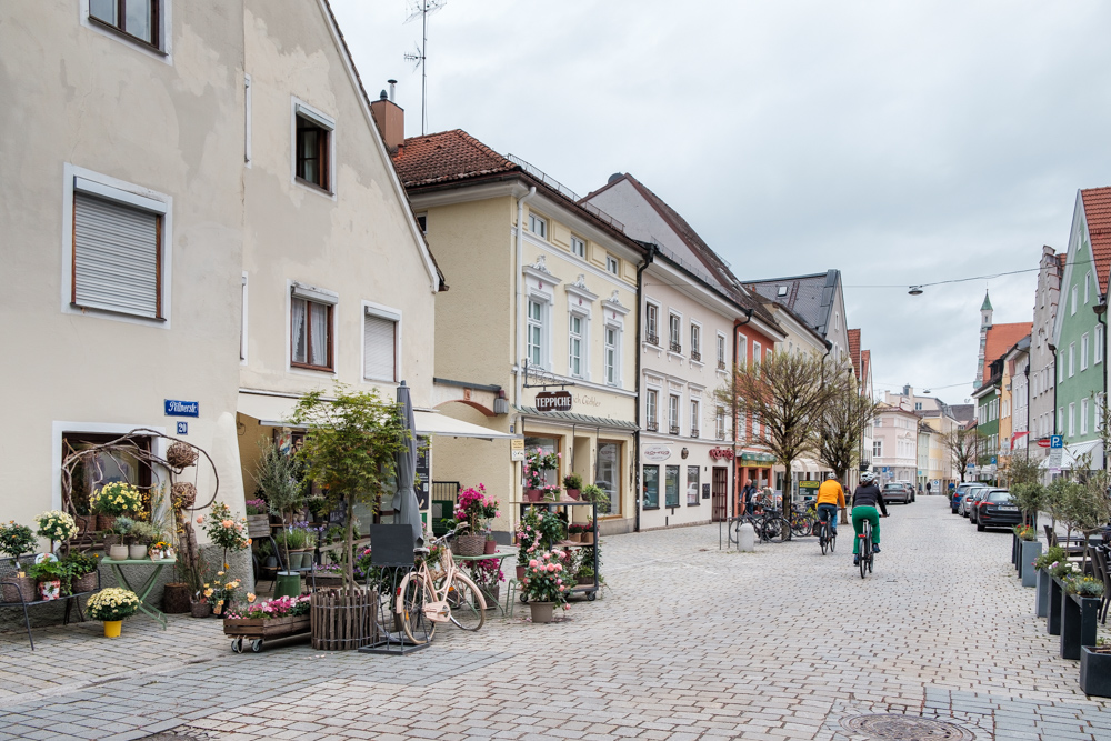 Zwei Fahrradfahrer auf einer gepflasterten Einkaufsstraße. Im Vordergrund: ein Blumenladen 