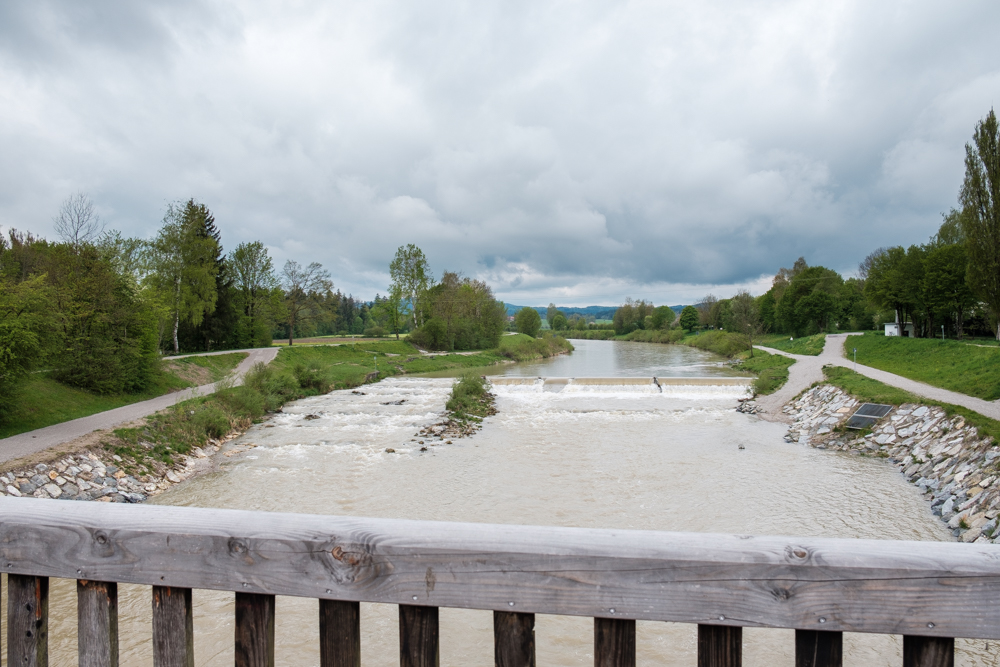 Foto gegen die Fließrichtung der Ammer. Im Vordergrund ein Holzgeländer der Brücke, auf dem die Fotografin steht. 